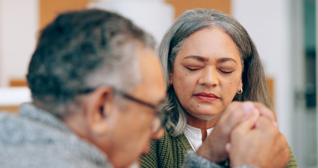 An image depicting an elderly couple, hand in hand, praying together at home, exemplifying 'prayer for married couples'. Their closed eyes and serene expressions convey hope, support, trust, and faith in their marriage commitment. This moment of bonding between the senior man and woman signifies their shared prayer for peace and worship.