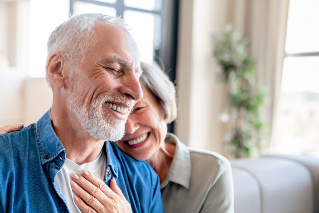 "A senior Caucasian couple demonstrating respect for husbands through physical touch, a love language. The elderly spouses are embracing in a warm hug at home, showcasing a moment of love, care, and companionship in their golden years."